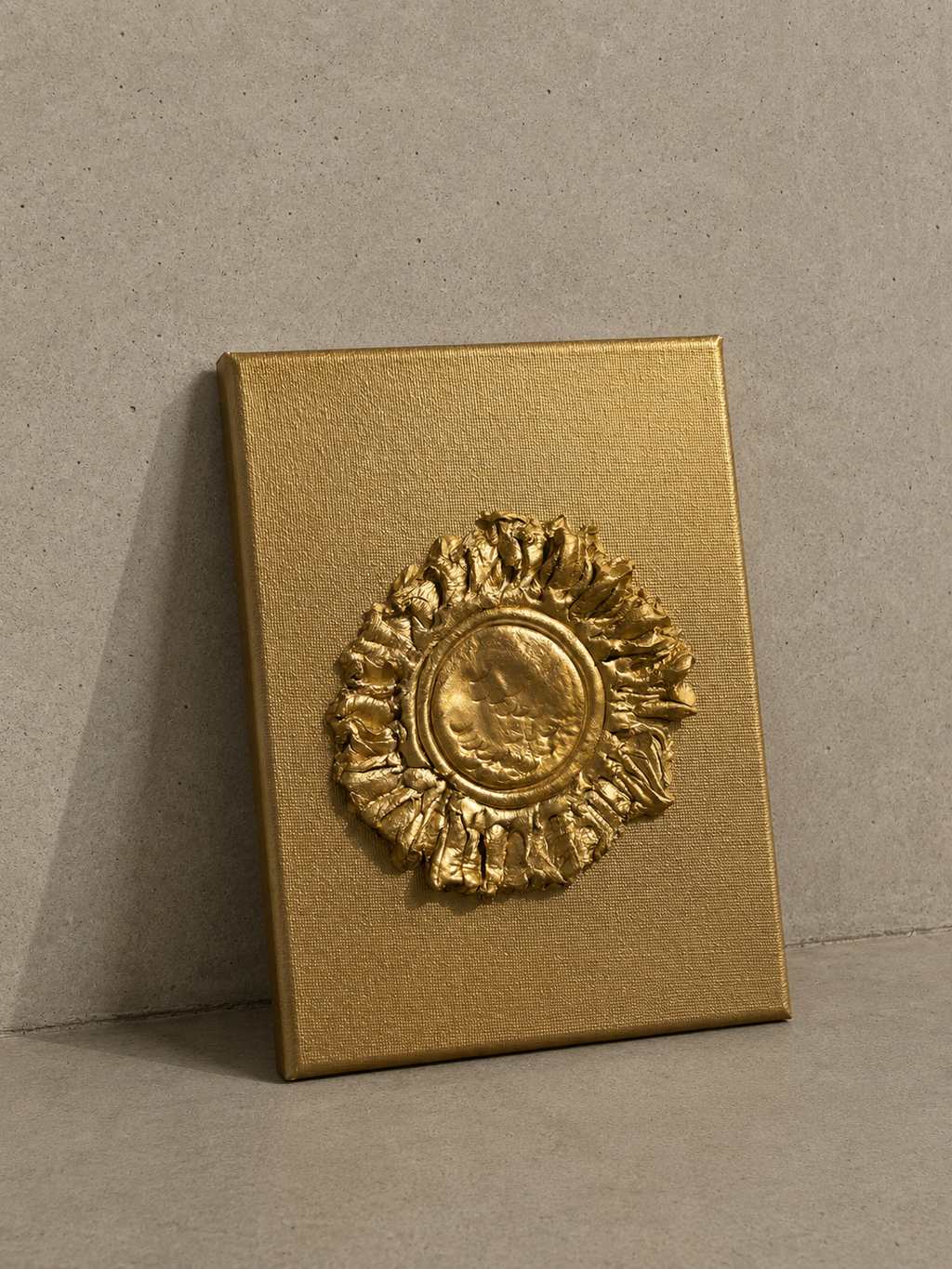 A square gold-toned plaque or box-like object with a raised circular sunburst medallion, photographed against a neutral textured gray wall and floor.