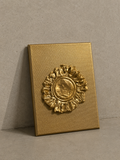 A square gold-toned plaque or box-like object with a raised circular sunburst medallion, photographed against a neutral textured gray wall and floor.
