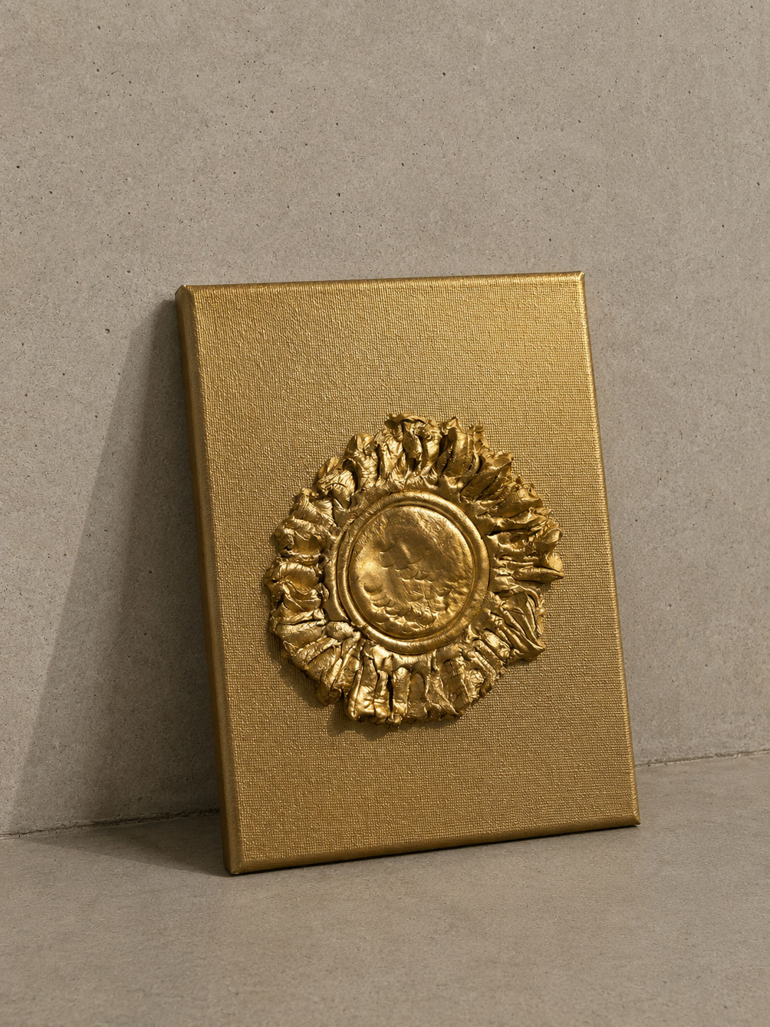 A square gold-toned plaque or box-like object with a raised circular sunburst medallion, photographed against a neutral textured gray wall and floor.