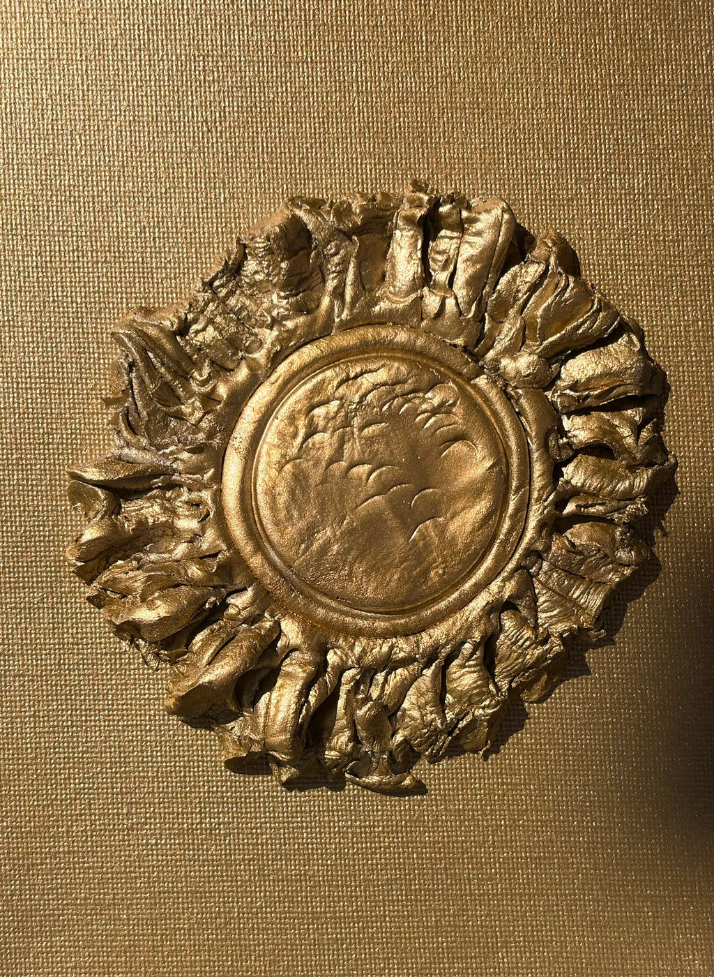 An ornate circular gold-toned decorative object with a textured central medallion and a sunburst-like ridged outer rim, shown against a warm tan woven fabric background.