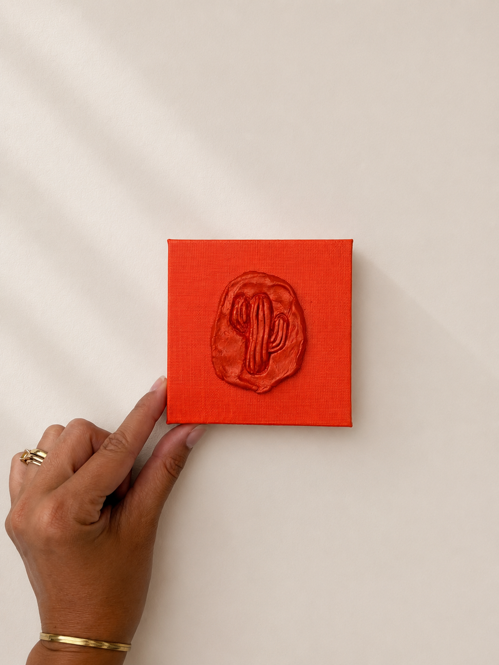 Cactus Arancia — a small orange ceramic cactus emblem mounted on a red box canvas, photographed held up against a plain white background.