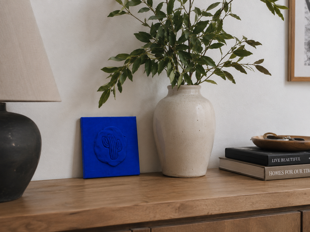 Cactus Blu — a small blue ceramic cactus emblem mounted on a red box canvas, photographed against a console table