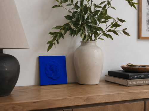 Cactus Blu — a small blue ceramic cactus emblem mounted on a red box canvas, photographed against a console table