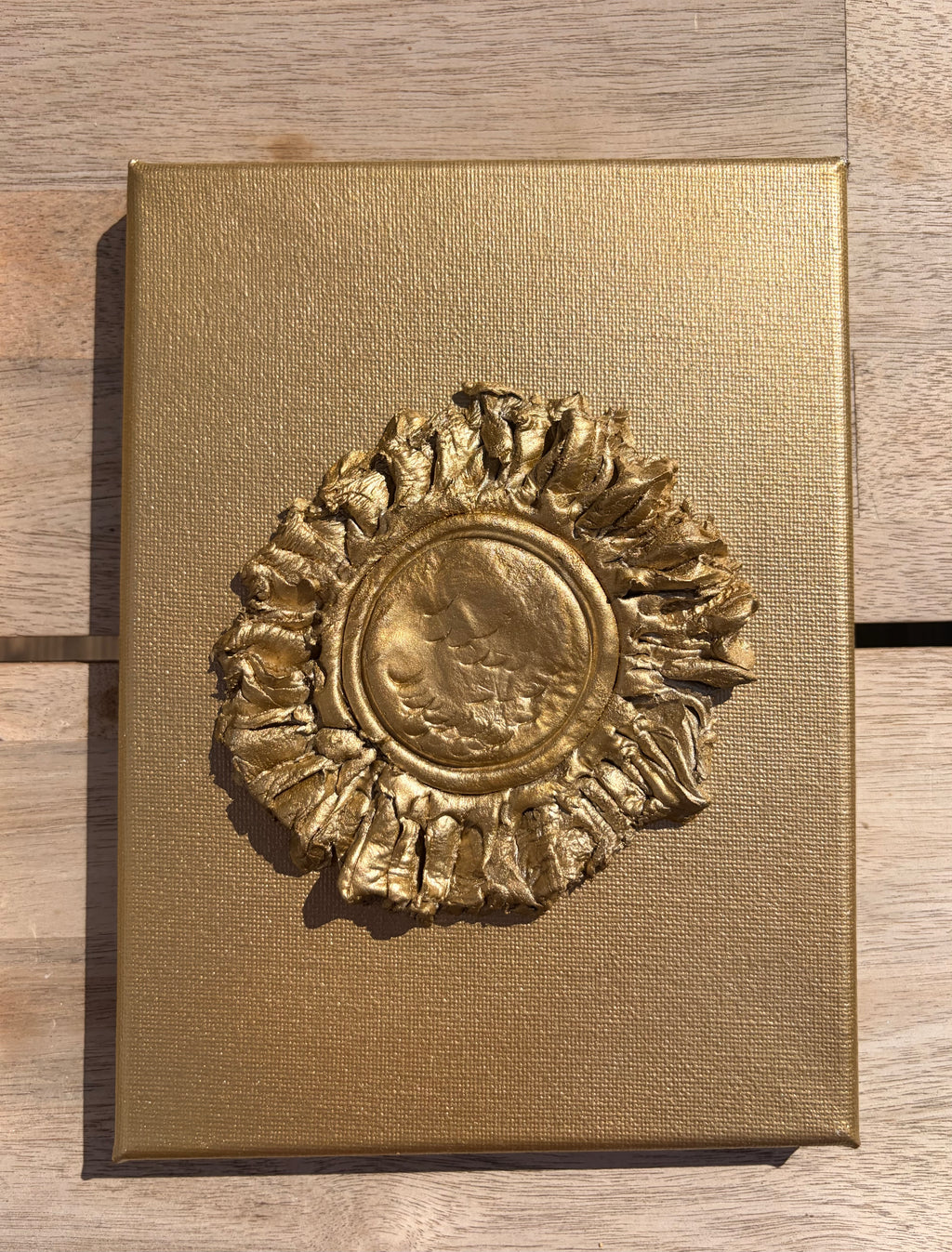 A square gold-toned metal plaque or decorative box lid featuring a raised textured emblem in the center, photographed from above on a light wood surface.