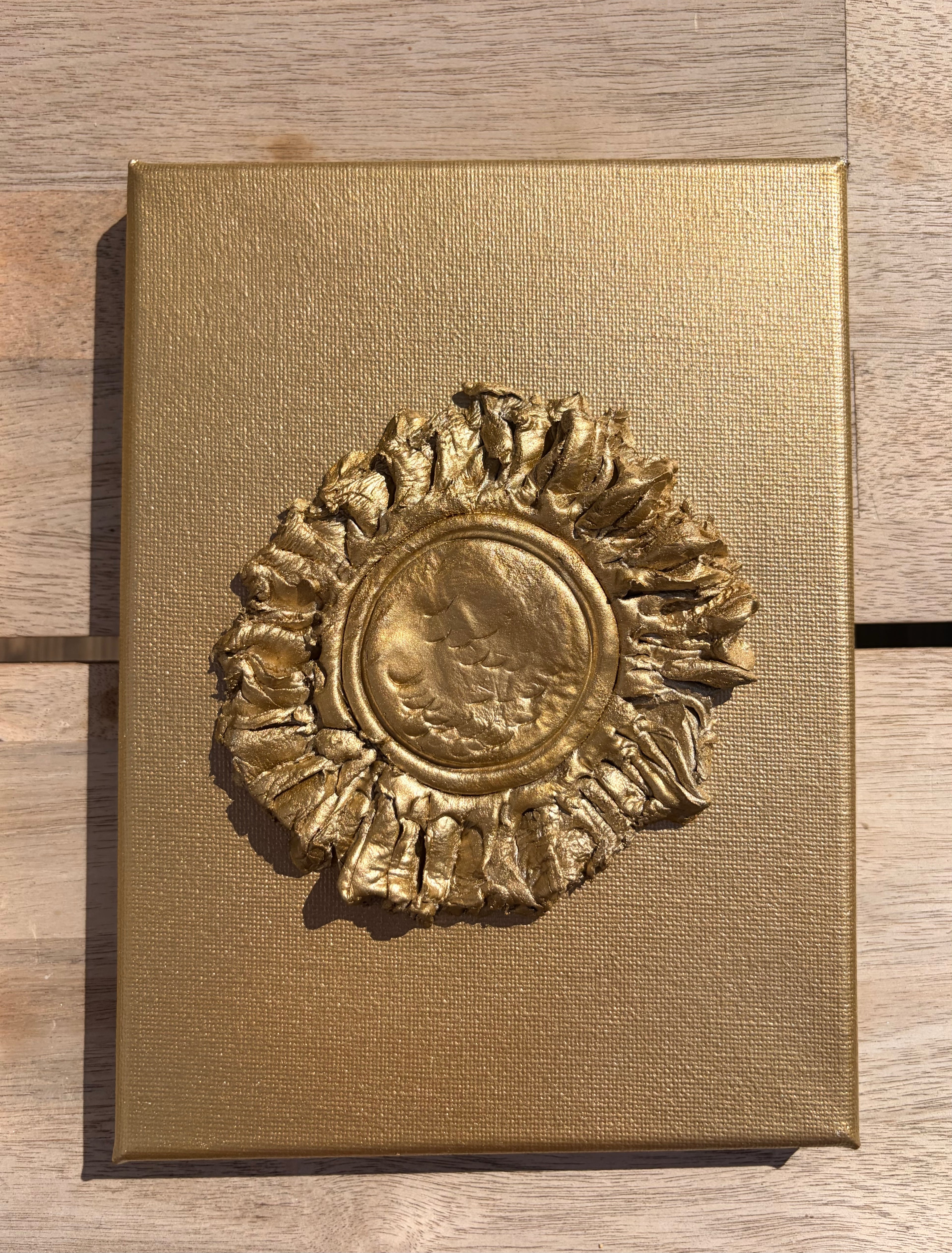 A square gold-toned metal plaque or decorative box lid featuring a raised textured emblem in the center, photographed from above on a light wood surface.